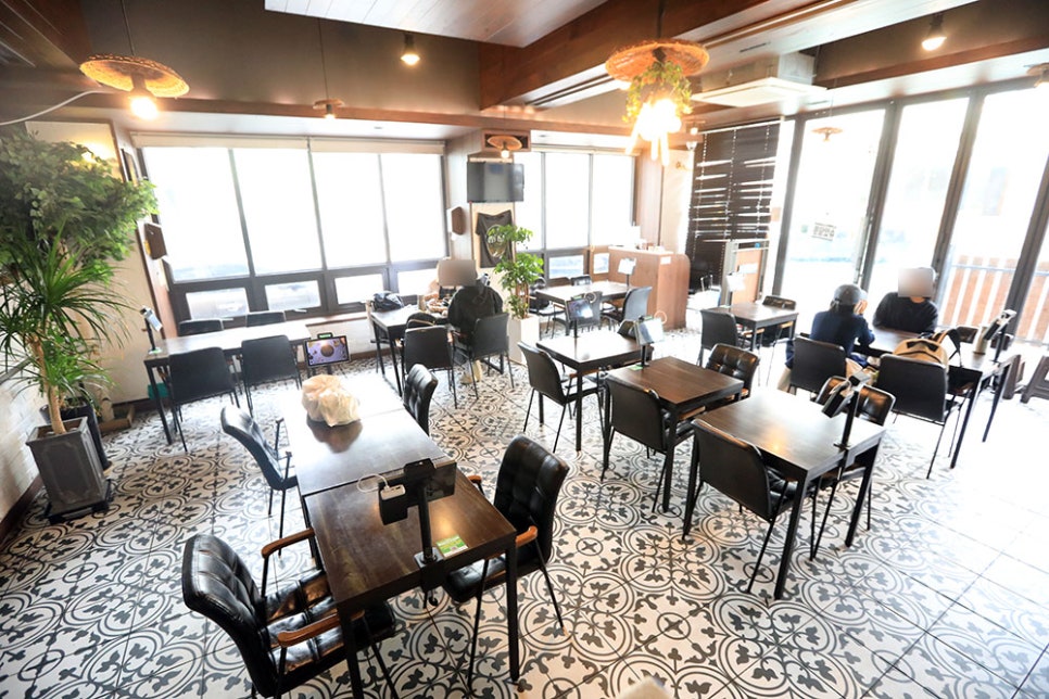 우드톤 천장과 푸릇한 화분 인테리어, 그리고 어두운 고동색 목제 테이블과 검정 의자로 구비돼 있는 가게 내부입니다. 