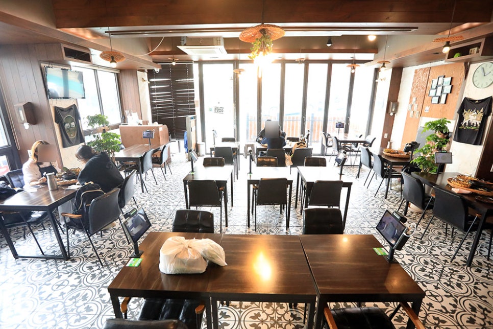 우드톤 천장과 푸릇한 화분 인테리어, 그리고 어두운 고동색 목제 테이블과 검정 의자로 구비돼 있는 가게 내부입니다. 